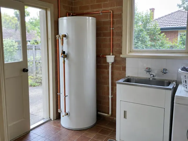 White hot water tank with copper pipework beside a laundry sink and window in Artarmon